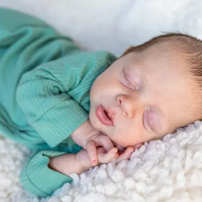 A newborn baby in a green ribbed onesie sleeping on a white textured blanket.