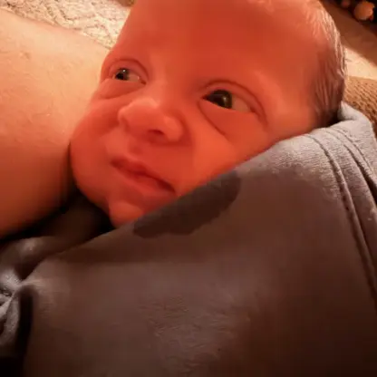 A newborn baby with a squinting expression nestled against an adults shoulder.