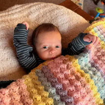 A newborn wearing a striped shirt lies on a fuzzy surface with their arms raised, partially covered by a pastel crocheted blanket.