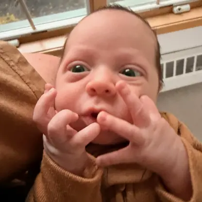 A newborn baby with wide eyes holds their hands near their face.