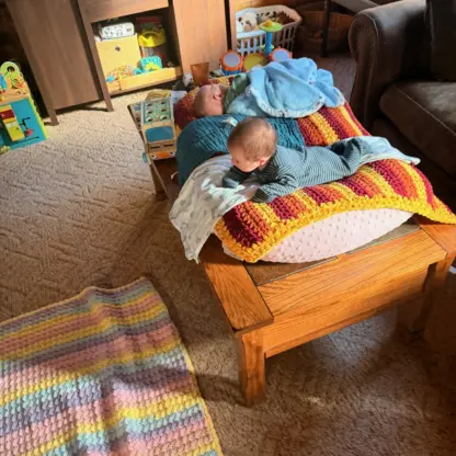 Two infants lie on a multi-colored crocheted blanket atop a wooden coffee table in a living room.