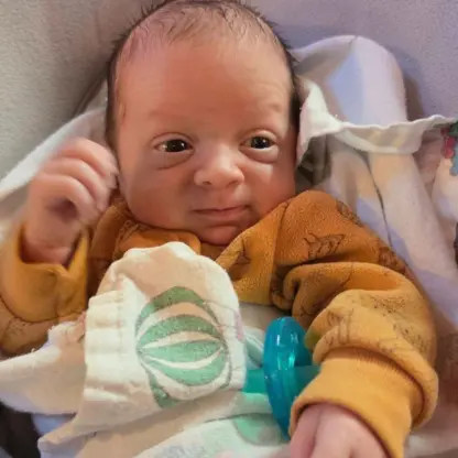 Newborn baby wearing a mustard-colored long-sleeve shirt and a pacifier, resting under a blanket.