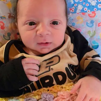 A newborn baby wearing a black and gold Purdue University shirt, resting on a colorful patterned blanket.