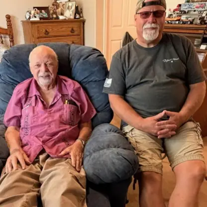 An elderly man sitting in a blue recliner next to a middle-aged man wearing a baseball cap and glasses.