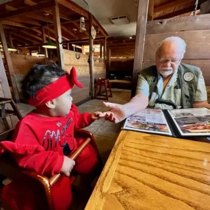 An older man in a vest sits at a wooden table in a restaurant, holding hands with a toddler in a red outfit who is sitting in a high chair.
