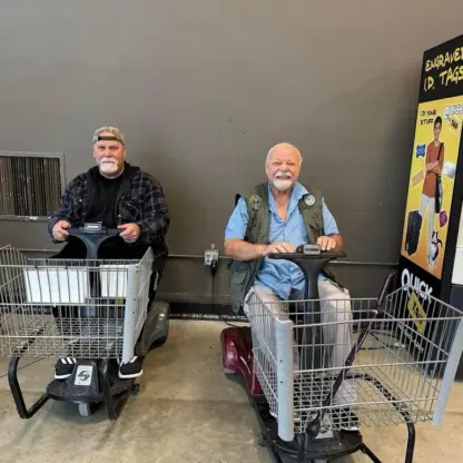 Two men sit on motorized shopping carts inside a store next to an engraved ID tag kiosk.
