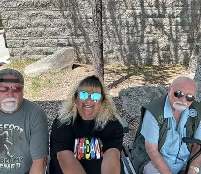Three people wearing sunglasses sit together on a bench outdoors.