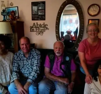 Four people sit together on a sofa in a living room, with an oval mirror and wall decorations behind them.