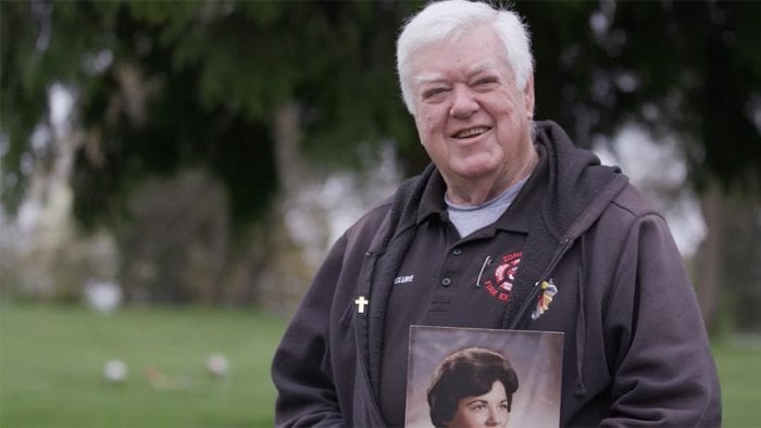 Elderly man with white hair holds framed photo of his deceased loved one in cemetery