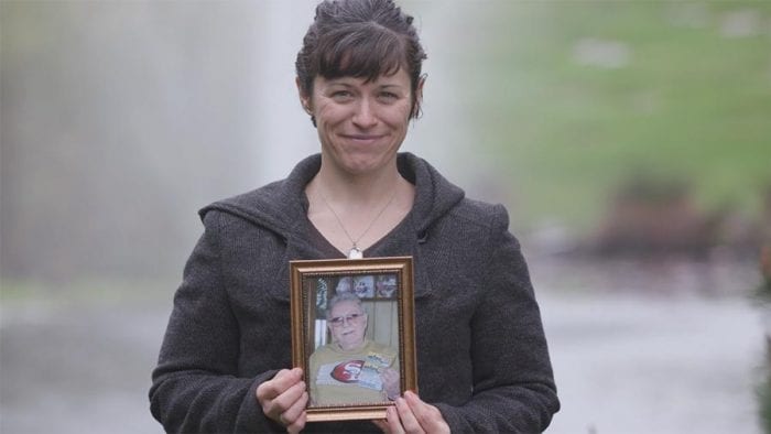 Woman smiles fondly in front of cemetery fountain holding framed photo of deceased loved one