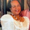 a woman wearing a white dress, beaded necklace, and glasses smiles