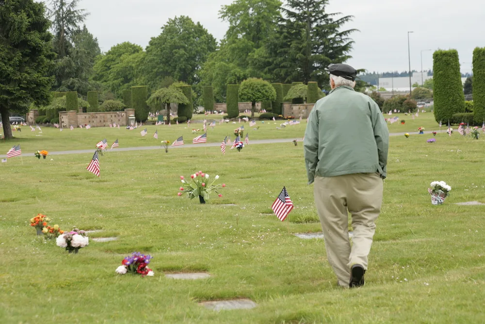 Older man in sage green jacket and khakis walks away in cemetery during Memorial Day celebration
