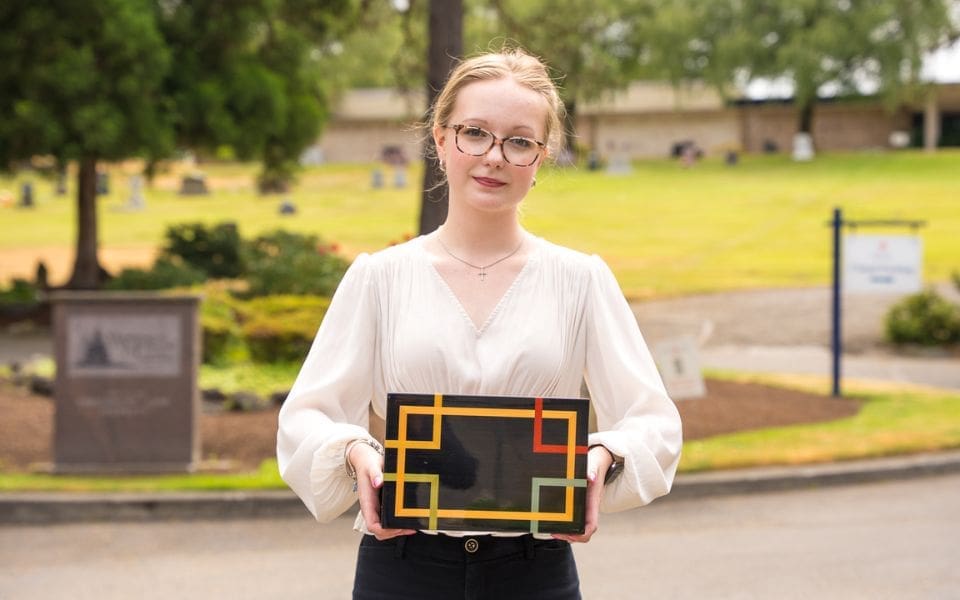 A young woman with glasses and a white blouse stands outside, holding a rectangular object with a black surface featuring Washington Memorial Park cemetery in the background.