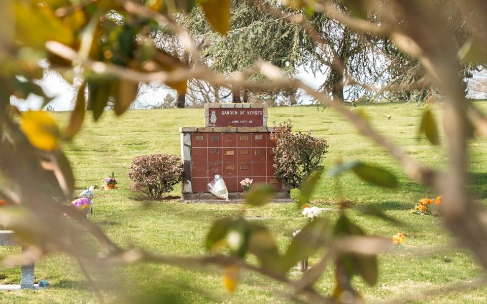 A grassy cemetery lawn at Washington Memorial Park, framed by blurred tree branches and leaves in the foreground. Colorful flower arrangements are scattered on the grass.