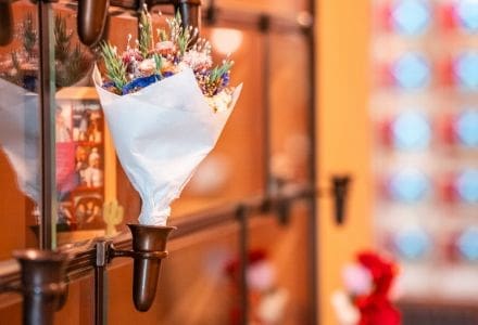 A small bouquet of flowers wrapped in white paper rests in a bronze holder attached to a glass-fronted columbarium niche, softly illuminated by colorful, blurred lights.
