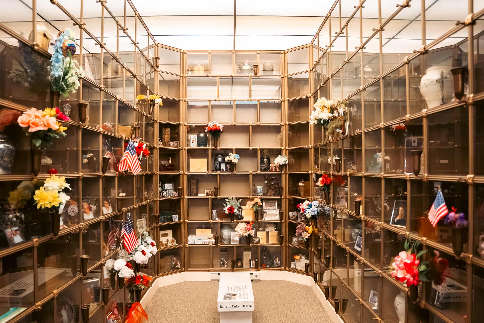 A mausoleum columbarium with glass-front niches containing urns, flowers, photos, and American flags for honoring loved ones. The serene walls offer a respectful atmosphere for remembrance, whether for pre-planning or in times of immediate need.