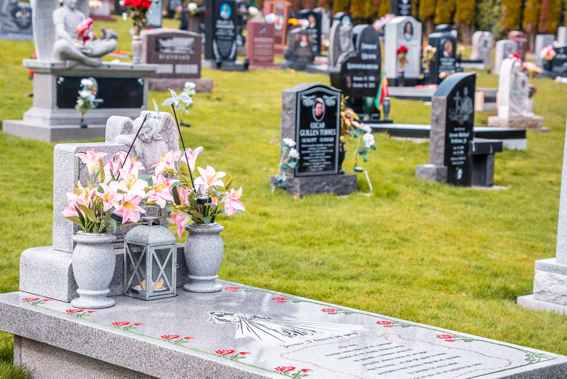 A cemetery with gravestones and headstones on a grassy lawn; one grave in the foreground is decorated with pink and white flowers in vases and a lantern. Other gravestones are visible in the background.