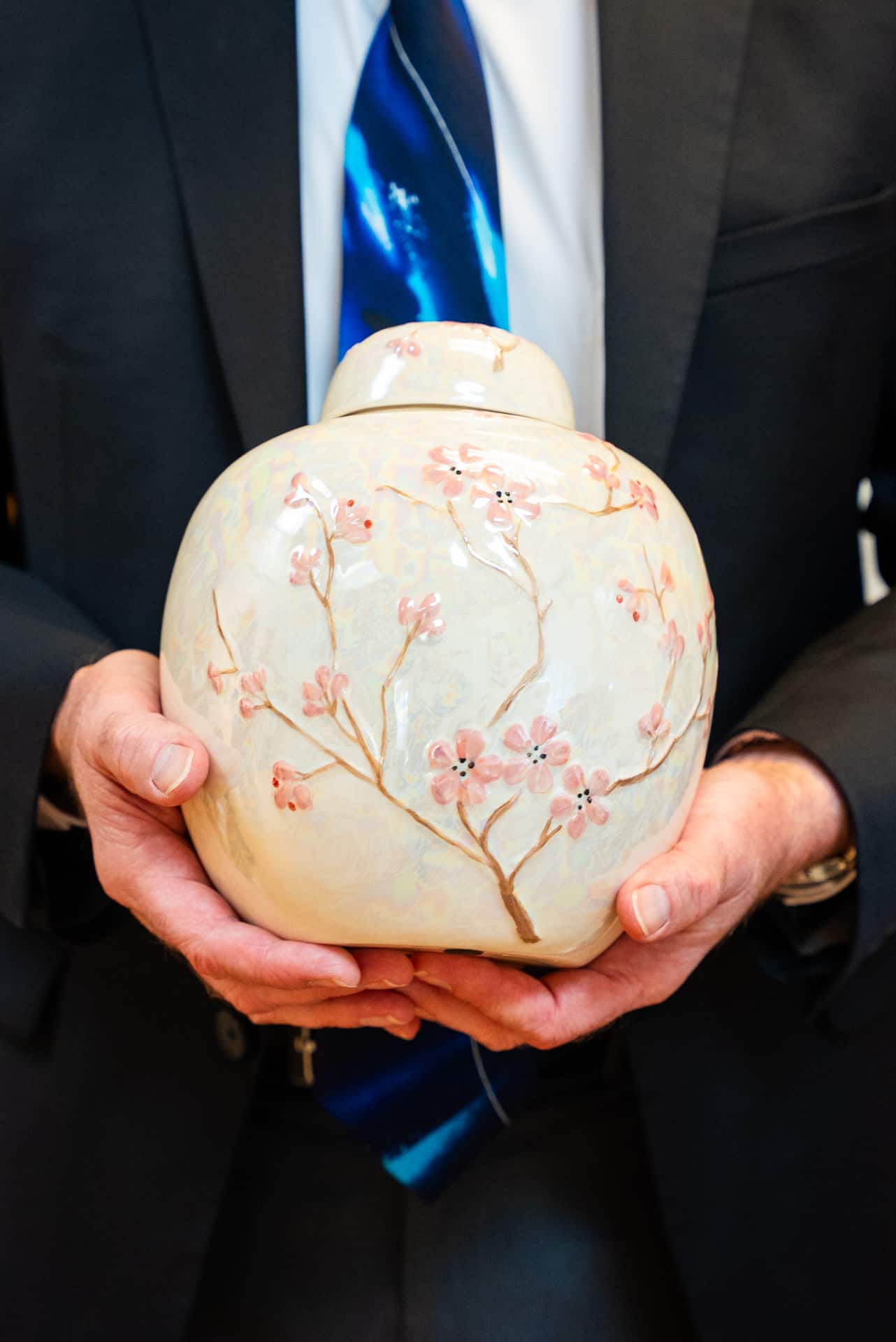 A person in a suit and blue tie gently holds a round ceramic urn, decorated with pink cherry blossom branches, at a Seattle funeral home.