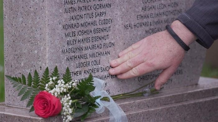 A person’s hand gently touches engraved names on a memorial stone, with a red rose and white flowers placed nearby in remembrance.