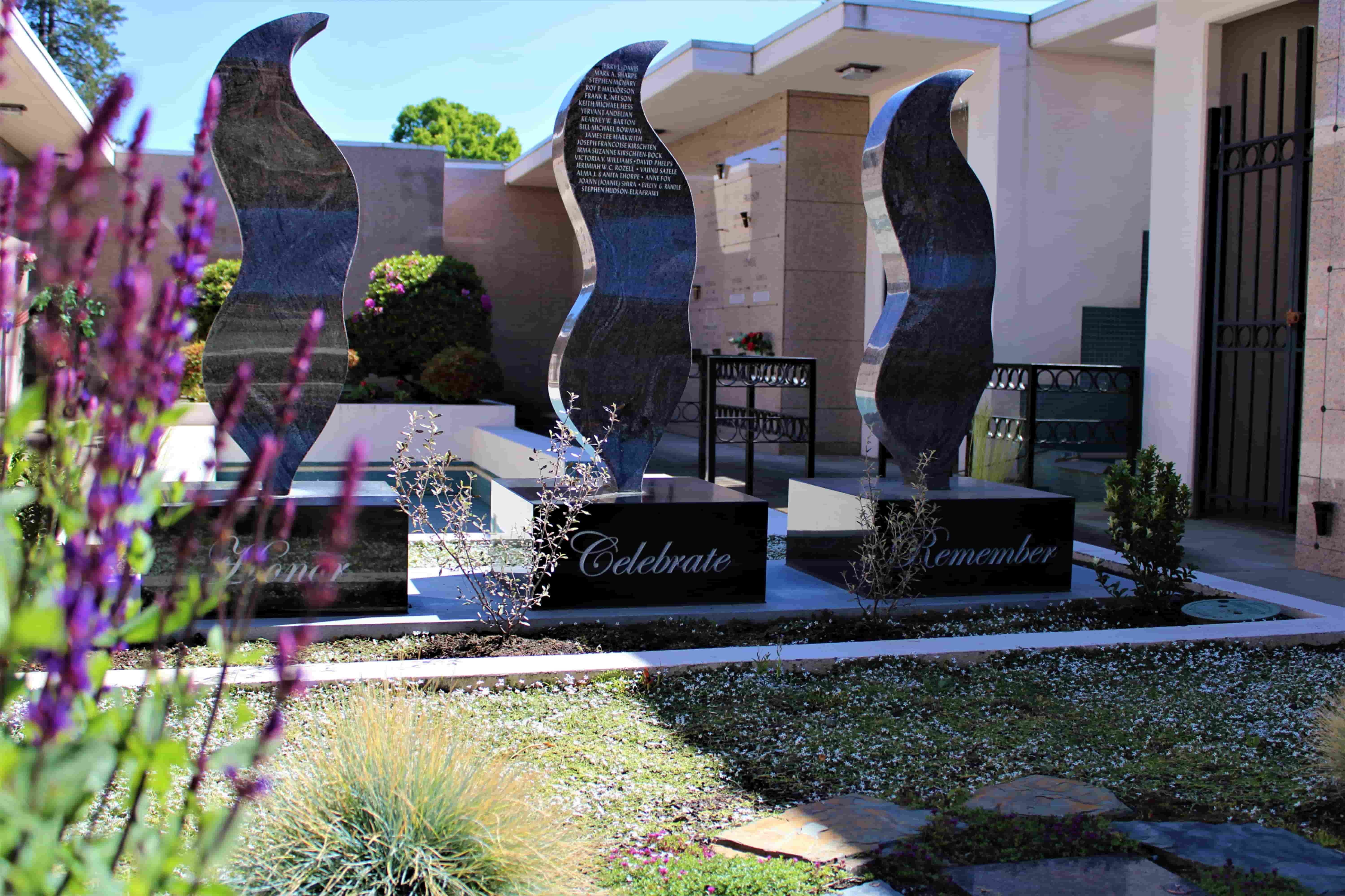 A memorial with three tall, flame-shaped stone sculptures stands outside a building offering funeral home services. The bases are inscribed with Love, Celebrate, and Remember, and the monument is surrounded by flowers and greenery.