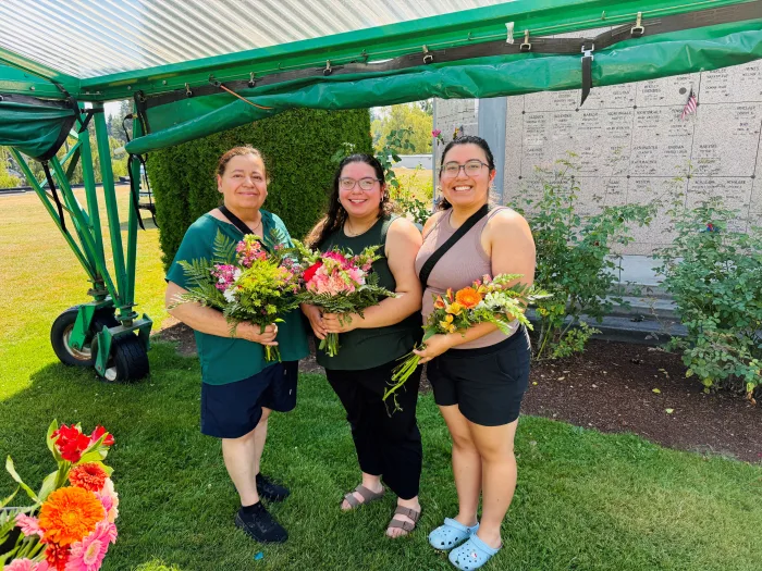 Three women stand outdoors under a green canopy, smiling and holding colorful bouquets of flowers. Behind them is a wall with plaques and green shrubbery on a sunny day.