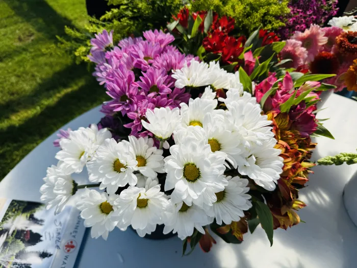 A bouquet of white daisies and purple chrysanthemums is arranged on a white table outdoors, with green grass and other flowers blurred in the background.