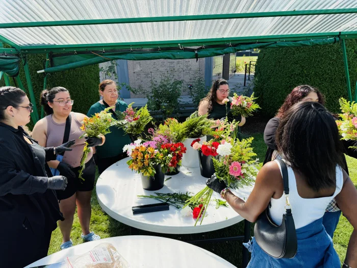 A group of people arranging colorful flower bouquets around white tables under a green canopy outdoors, with various flowers and greenery placed in black vases.