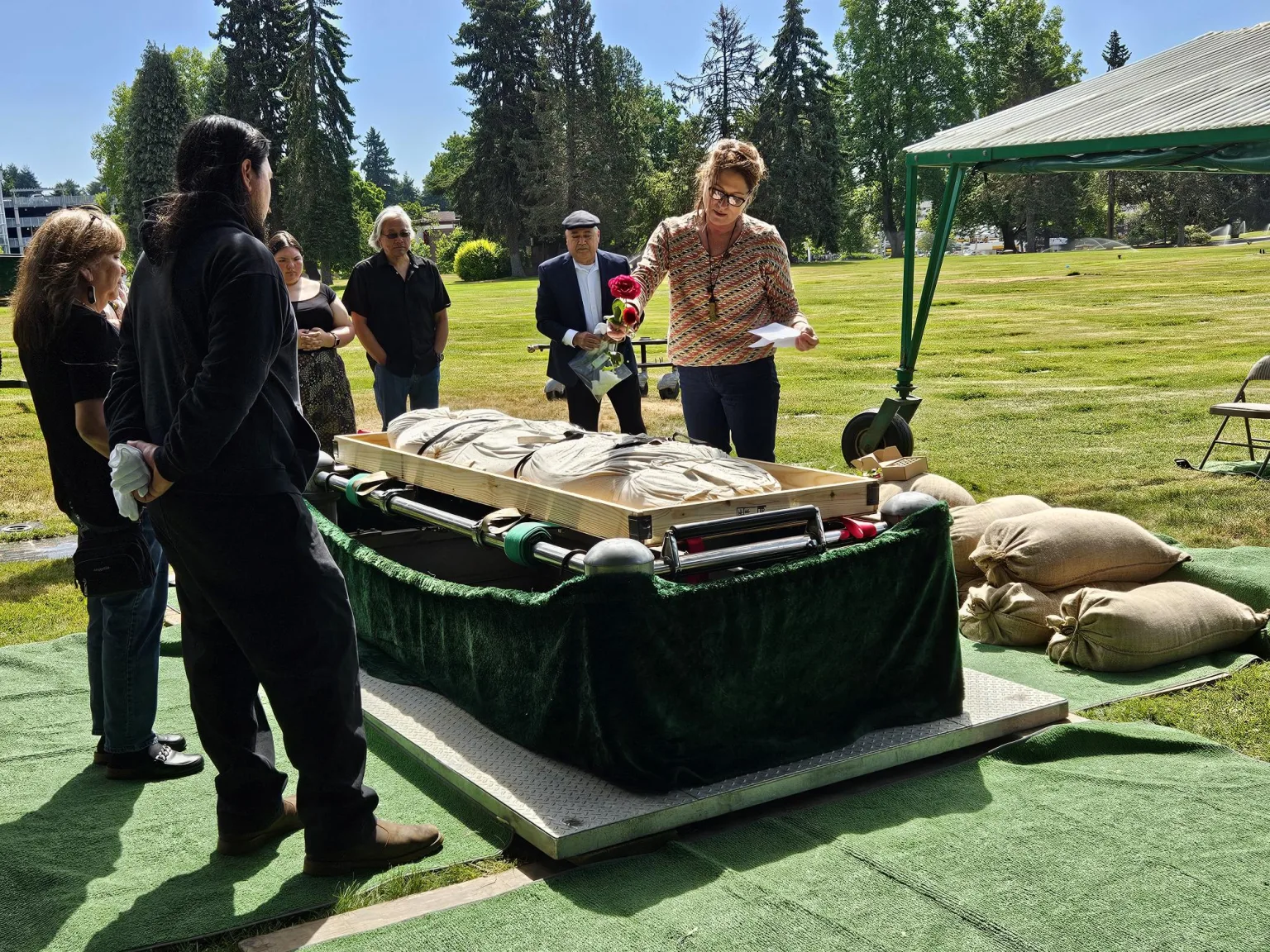 A group of people stand around at a burial service in a cemetery. One person, honoring the terramation process, holds a red rose and reads from a piece of paper to the others under a green canopy.