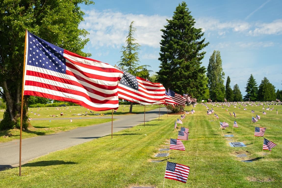 Large American flags line a road at Washington Memorial Park cemetery, with smaller flags on graves throughout the grassy area. Tall trees and a clear blue sky create a peaceful scene, honoring loved ones in this resting place that supports terramation and natural remembrance.