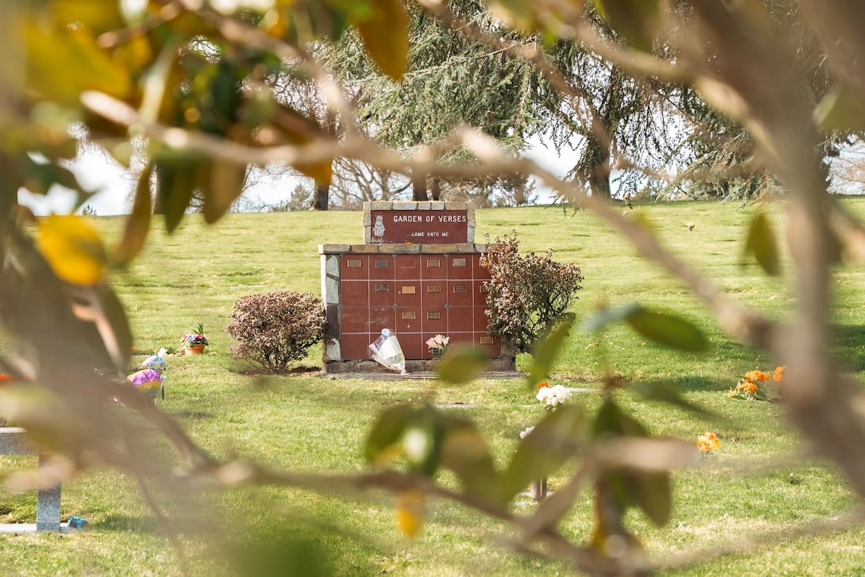 El «Jardín de los Recuerdos» se encuentra en un cementerio que ofrece servicios funerarios, rodeado de césped, pequeños arbustos y flores dispersas, con las ramas de los árboles enmarcando parcialmente la vista.