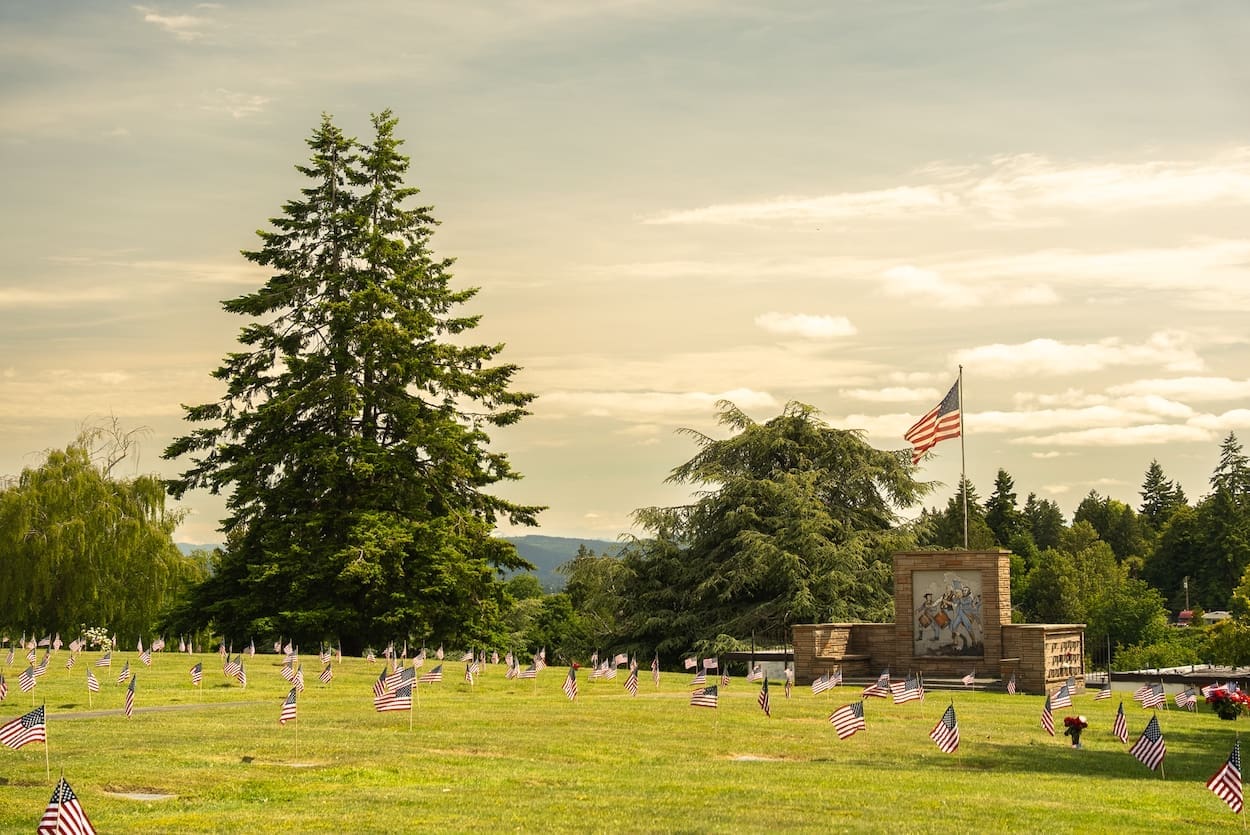 A cemetery lawn is dotted with numerous small American flags, honoring those remembered through veterans services. In the background, tall trees and a memorial wall with an American flag flying above it are visible under a partly cloudy sky.