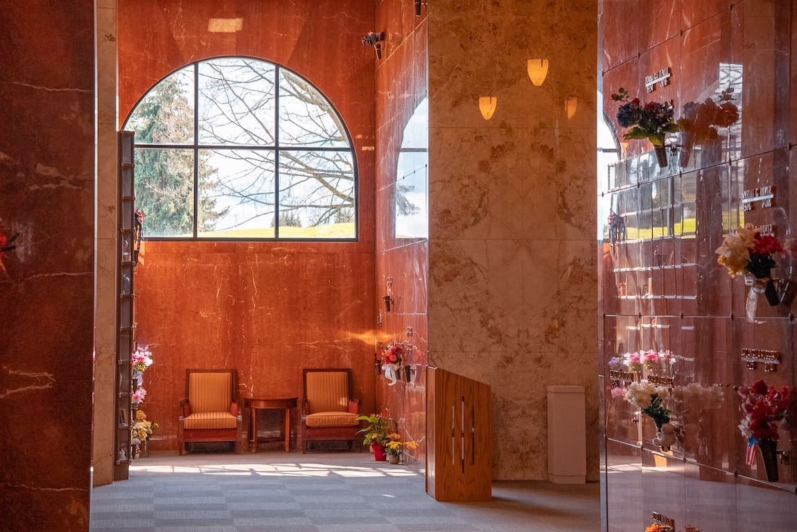 El tranquilo interior del mausoleo de BONNEY WATSON, con paredes de mármol, arreglos florales en las criptas, dos sillas junto a un gran ventanal en arco y una suave luz natural que inunda el espacio.