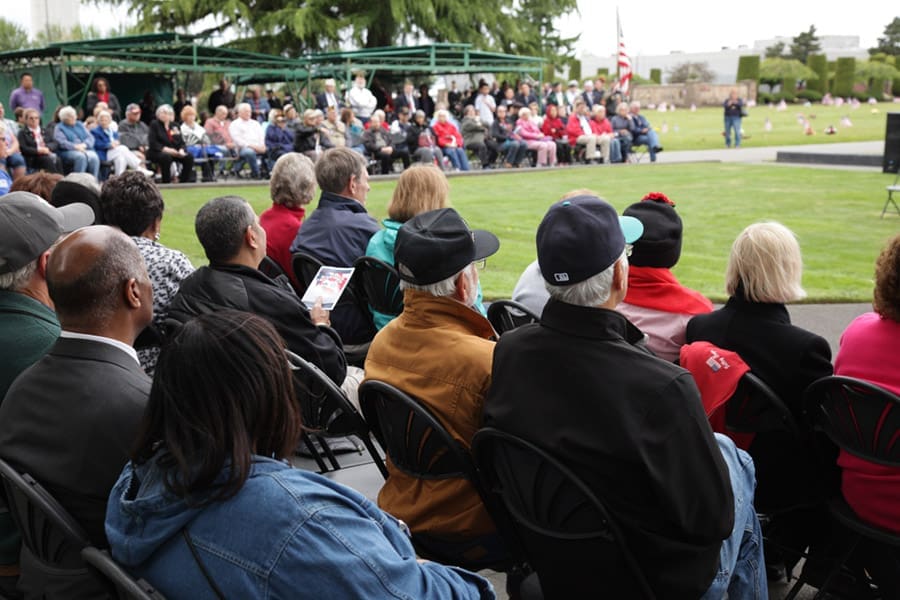 A diverse group of people sit on black chairs outdoors, attentively watching a ceremony on a grassy area. More attendees and a U.S. flag can be seen in the background beneath cloudy skies.