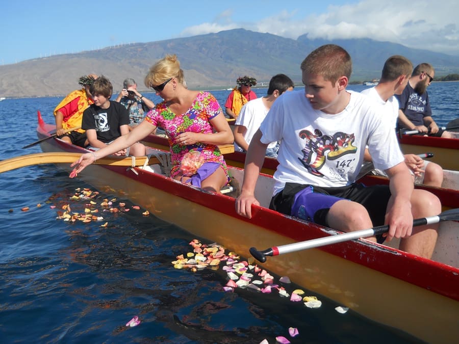 People in two canoes on a calm body of water scatter colorful flower petals during a ceremony. A mountain and blue sky are visible in the background. The group includes adults and teenagers.