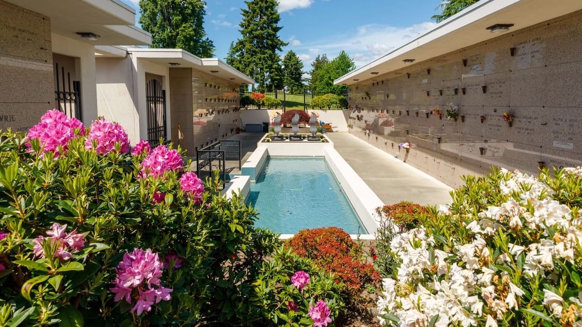 A peaceful outdoor mausoleum with flower arrangements, green shrubs, and blooming pink and white flowers surrounding a central reflecting pool under a bright blue sky.