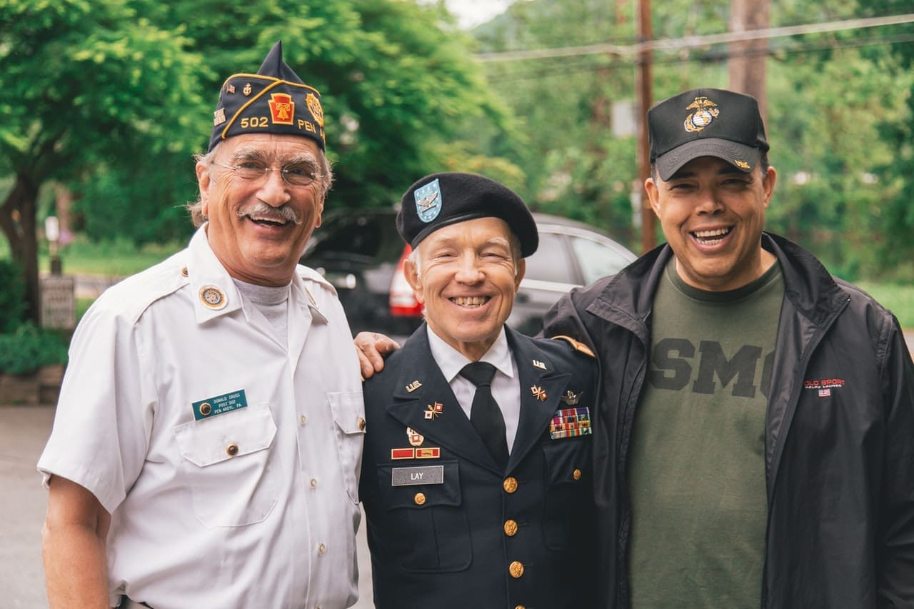 Three smiling men stand together outdoors. Two wear military uniforms and hats, while the third wears a black jacket and cap. Enjoying the moment, they appear happy and relaxed, perhaps celebrating veterans services with green trees and a street behind them.