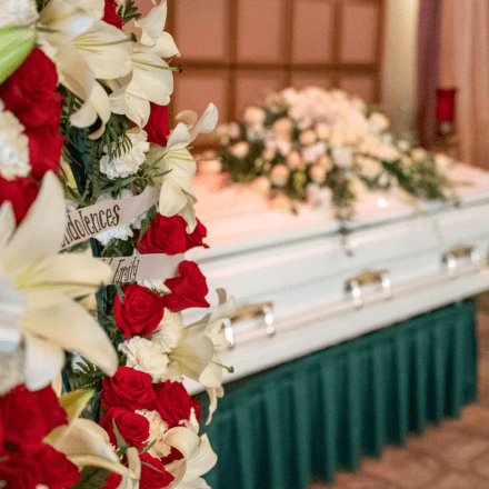 A white casket with a flower arrangement rests at the front of a room; in the foreground, a funeral wreath with red and white flowers and a ribbon reading Condolences is visible, reflecting thoughtful selections for every need and pricing.