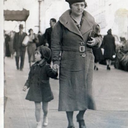 A woman in a long coat and hat walks hand-in-hand with a young girl in a coat and bonnet on a busy city sidewalk, with people and buildings in the background.