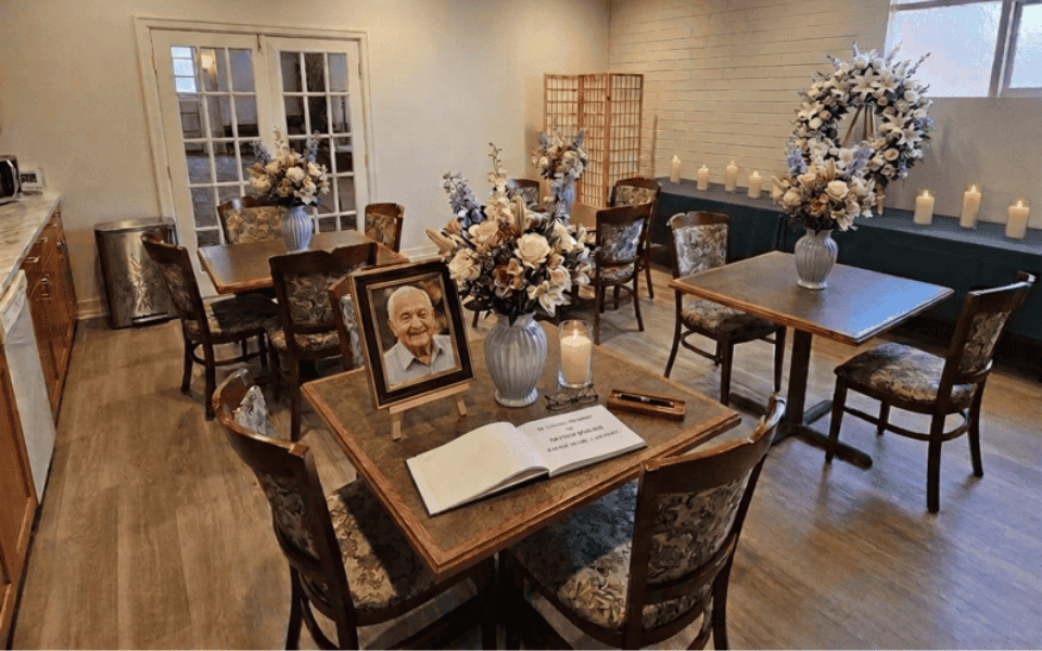 A memorial room designed for funeral planning, with floral arrangements, a framed photo of an elderly man, a guestbook on a table, candles on a counter, and several wooden tables and chairs. The atmosphere is calm and respectful.