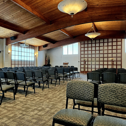 Rows of empty chairs arranged in a spacious room with wooden ceiling beams, carpeted floor, and soft overhead lighting create a calm atmosphere suitable for funeral planning. A stage with a podium and U.S. flag is in the background.