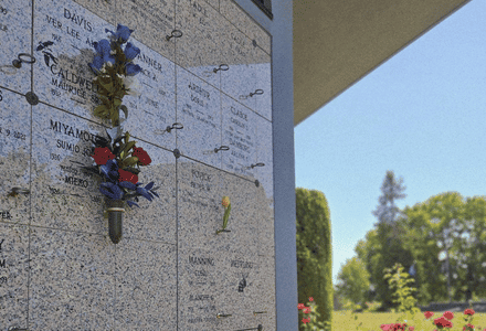 A marble columbarium wall with engraved nameplates, metal flower holders, and colorful flowers offers a serene setting ideal for thoughtful funeral planning. Green trees, grass, and a blue sky are visible in the background.