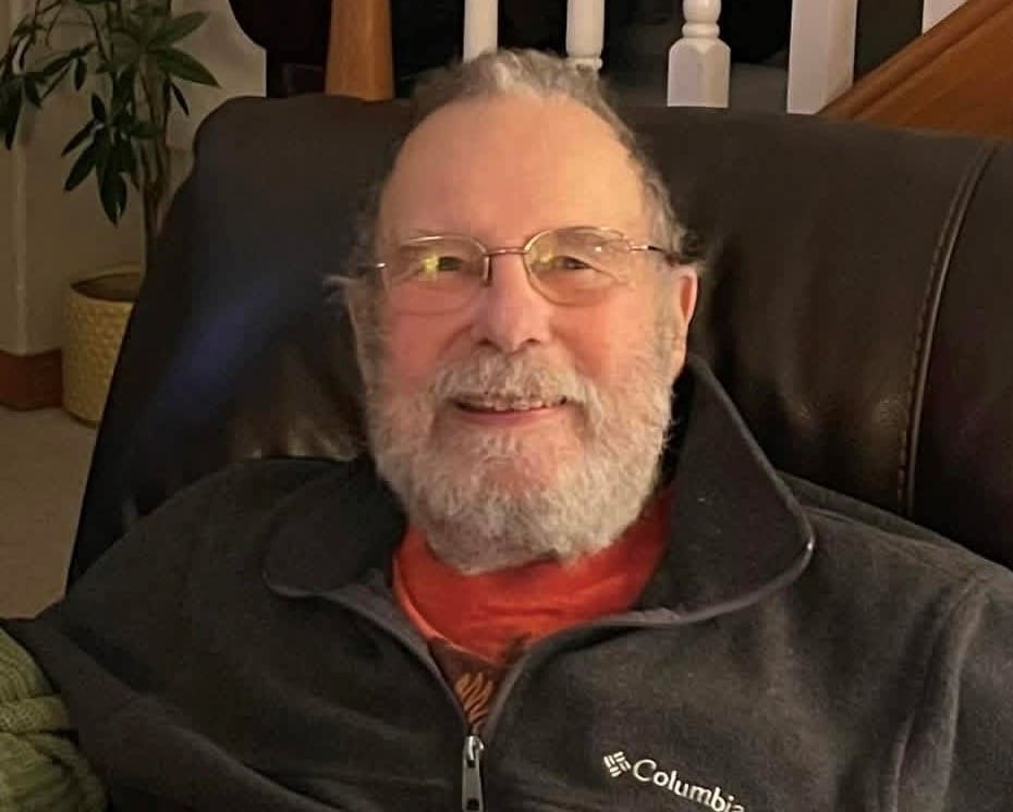 An older man with glasses and a gray beard smiles while sitting on a dark chair, wearing a red shirt and a gray Columbia jacket. A plant and staircase are in the background.